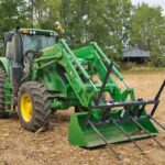 A John Deere tractor and bush-hogging attachment sit in a freshly cut corn field with an old barn in the background.